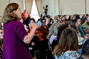 Candid photograph of a seated crowd focused on a woman standing in the foreground. The woman is mid-speech, gesturing with her right arm in front of her chest.