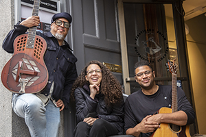 Photograph of three people sitting outside of Mechanics' Hall. From left to right: Samuel James, a broad masculine presenting person with brown skin, a grey beard, and a dark jean jacket holding a guitar with a unique geometric pattern on his right knee; Maya Williams, a feminine presenting person with brown skin, tight curled hair that ends around their shoulder blades, and a broad open-mouthed smile; Brian Evans, a masculine presenting person with short curly hair, a wide close-lipped smile, and wearing a black long-sleeved shirt, holding a traditional acoustic guitar.