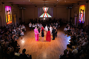 Photograph from the opening night of Carmen. Featuring a circle of performers holding hands surrounded by audience members, hosted in Mechanics' Hall Ballroom.