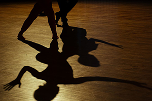 Photograph of a Little House Dance performance, focused on the silhouette of two dancers' legs at the top of the photo and their full shadows casted on a wooden floor.