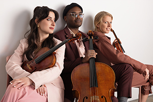 Photo of three members from Palaver Strings posing with their instruments. From left to right: A white woman with long dark hair and a full-face of makeup and looking away from the camera while hugging a viola on her lap, a Black man wearing glasses and a maroon suit staring towards the camera while holding the neck of a cello, and a white woman seated facing away from the camera looking over her shoulder towards the camera while holding a violin on her lap.